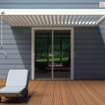 Nowoczesna biała pergola lamelowa Marlo zamontowana na tarasie przy budynku mieszkalnym. Konstrukcja pergoli opiera się na dwóch pionowych, kwadratowych słupach z przodu oraz jest przytwierdzona bezpośrednio do elewacji budynku, która wykończona jest poziomym sidingiem w kolorze szarym. Zadaszenie składa się z ruchomych, równolegle ułożonych białych lameli, które są obecnie ustawione pod kątem, pozwalając na częściowy dopływ światła słonecznego. Po lewej stronie konstrukcji widoczna jest zwisająca biała korba służąca do ręcznej regulacji kąta nachylenia lameli. Pod pergolą znajduje się taras wykonany z desek kompozytowych lub drewnianych w naturalnym, miodowym odcieniu, ułożonych prostopadle do elewacji. Na tarasie ustawiony jest ciemnobrązowy leżak z jasnoszarą poduszką oraz mały, niski stolik kawowy. W tle widać duże, przeszklone drzwi przesuwne prowadzące do wnętrza domu, w których odbija się zieleń ogrodu. Cień rzucany przez pergolę na elewację i podłogę tarasu jest wyraźny, co wskazuje na słoneczną pogodę.
