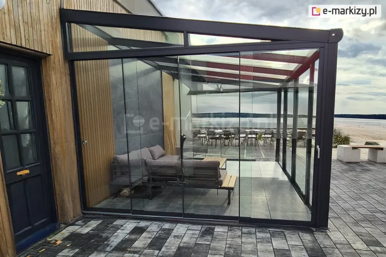 Taras wypoczynkowy osłonięty przed wiatrem za pomocą szklanych paneli przesuwnych Auris. Konstrukcja tworzy przezroczystą, lekką wizualnie barierę, która pozwala korzystać z tarasu niezależnie od warunków pogodowych. Wzdłuż krawędzi tarasu zamontowane są aluminiowe prowadnice, po których przesuwają się duże tafle szkła, a same panele tworzą ciągłą osłonę boczną. We wnętrzu strefy wypoczynkowej widoczne są meble ogrodowe, w tym niska sofa i stolik. Szkło zapewnia niezakłócony widok na otoczenie, w tym przestrzeń zastawioną stolikami i krzesłami, oraz ławeczkę przy piaszczystej plaży na brzegu jeziora, a jednocześnie chroni przestrzeń tarasu przed podmuchami wiatru. Całość konstrukcji prezentuje się nowocześnie i minimalistycznie, podkreślając charakter eleganckiej, funkcjonalnej zabudowy tarasowej z użyciem systemu Auris.