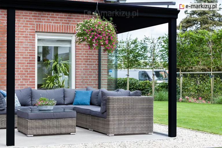 Narożna część zadaszenia tarasu Terano, zamontowanego do elewacji z czerwonej cegły. Konstrukcja aluminiowa w kolorze antracytowym opiera się na solidnych, kwadratowych słupach nośnych. Pod zadaszeniem znajduje się strefa relaksu wyposażona w narożną sofę z technorattanu w kolorze szarym z ciemniejszymi poduszkami siedziskowymi oraz dopasowany stolik kawowy z szklanym blatem. Do konstrukcji zadaszenia przymocowana jest wisząca donica z obficie kwitnącymi, różowymi kwiatami, co nadaje przestrzeni przytulny charakter. Taras wyłożony jest jasnymi płytami wielkoformatowymi, a jego krawędź wykończona jest opaską z jasnego grysiku. W tle, za szklanym systemem przesuwnym, widoczny jest zadbany ogród z zielonym trawnikiem, żywopłotem oraz młodymi drzewami, a w oddali biały samochód dostawczy.