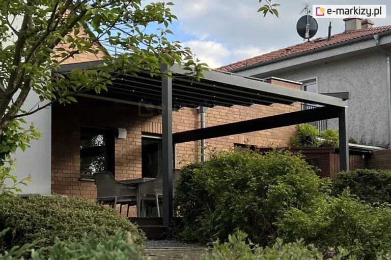 Aluminiowa pergola tarasowa Square w kolorze ciemnego antracytu, zamontowana do ściany budynku wykonanej z czerwonej cegły klinkierowej. Konstrukcja zadaszenia opiera się na poprzecznych profilach, na których rozpięta jest jasna, kremowa tkanina układająca się w regularne pasy. W profilach zadaszenia zamontowano punktowe oświetlenie LED, które rzuca delikatne światło na przestrzeń poniżej. Całość wspiera się na solidnym, pionowym słupie o kwadratowym przekroju, a z boku widoczna jest pozioma belka wzmacniająca konstrukcję. Pod pergolą, na podwyższonym tarasie, znajduje się zestaw mebli ogrodowych, w tym krzesła z szarej plecionki ustawione przy stole. Taras jest otoczony gęstą, naturalną roślinnością – na pierwszym planie oraz po prawej stronie widoczne są rozłożyste, zielone krzewy, które częściowo przesłaniają widok na meble. W górnej części zdjęcia zwieszają się gałęzie drzewa liściastego z wyraźnie zarysowanymi zielonymi liśćmi. W tle widać fragment sąsiedniego piętrowego domu o jasnej elewacji z ciemnoczerwonym dachem, na którym zamontowana jest antena satelitarna. Niebo jest zachmurzone, w odcieniach szarości i błękitu, co nadaje zdjęciu naturalny, dzienny charakter.