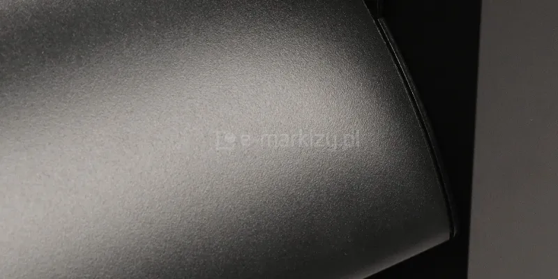 Zdjęcie prezentuje bardzo bliskie ujęcie powierzchni aluminiowego panelu żaluzjowego Louvre do pergoli w kolorze antracytowym. Widoczna jest drobnoziarnista, matowa struktura lakieru proszkowego, która nadaje elementowi elegancki, techniczny wygląd. Na zaokrąglonej krawędzi lameli skupia się miękkie światło, tworząc delikatny gradient i podkreślając satynowe wykończenie materiału. Po prawej stronie kadru widoczny jest fragment pionowego profilu konstrukcyjnego, co ukazuje precyzję spasowania poszczególnych elementów systemu louvre z konstrukcją pergoli lub zadaszenia. Kolorystyka jest jednolita, utrzymana w ciemnych odcieniach szarości, typowych dla nowoczesnej architektury ogrodowej.