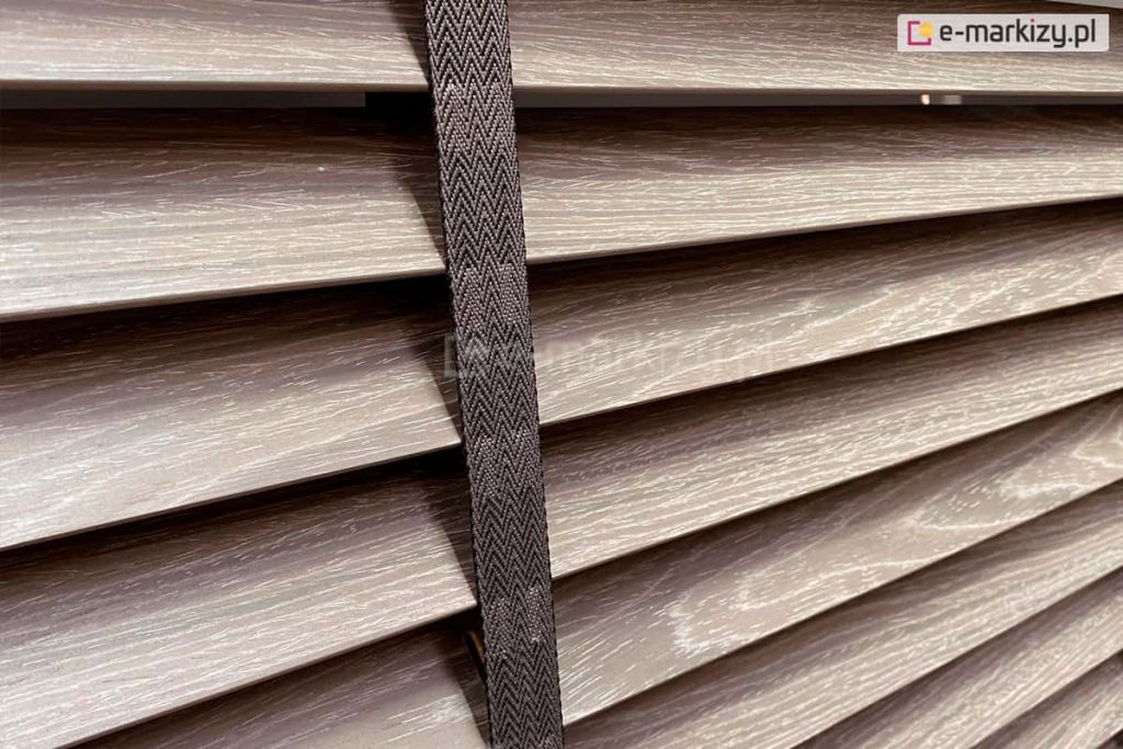 Zdjęcie prezentuje zbliżenie na żaluzje drewniane 50 mm w nowoczesnym, chłodnym odcieniu szarobrązowym z wyraźnie zarysowaną teksturą naturalnego usłojenia drewna. Centralnym punktem fotografii jest pionowa drabinka taśmowa w kolorze ciemnoszarym lub grafitowym, wykonana z gęstego splotu tekstylnego o wzorze jodełki. Taśma przebiega pionowo przez środek kadru, estetycznie przykrywając miejsca, w których żaluzje drewniane 50 mm posiadają otwory na sznurki prowadzące. Poszczególne lamele są ustawione pod lekkim kątem, co pozwala zaobserwować ich matowe wykończenie oraz precyzyjne krawędzie. Światło padające na powierzchnię drewna podkreśla białe akcenty w strukturze słojów, nadając osłonie surowy, industrialny charakter. Konstrukcja ukazuje wariant wykończenia premium, gdzie szeroka taśma pełni funkcję zarówno dekoracyjną, jak i użytkową, eliminując prześwity światła w miejscach perforacji lameli.