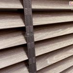 Zdjęcie prezentuje zbliżenie na żaluzje drewniane 50 mm w nowoczesnym, chłodnym odcieniu szarobrązowym z wyraźnie zarysowaną teksturą naturalnego usłojenia drewna. Centralnym punktem fotografii jest pionowa drabinka taśmowa w kolorze ciemnoszarym lub grafitowym, wykonana z gęstego splotu tekstylnego o wzorze jodełki. Taśma przebiega pionowo przez środek kadru, estetycznie przykrywając miejsca, w których żaluzje drewniane 50 mm posiadają otwory na sznurki prowadzące. Poszczególne lamele są ustawione pod lekkim kątem, co pozwala zaobserwować ich matowe wykończenie oraz precyzyjne krawędzie. Światło padające na powierzchnię drewna podkreśla białe akcenty w strukturze słojów, nadając osłonie surowy, industrialny charakter. Konstrukcja ukazuje wariant wykończenia premium, gdzie szeroka taśma pełni funkcję zarówno dekoracyjną, jak i użytkową, eliminując prześwity światła w miejscach perforacji lameli.