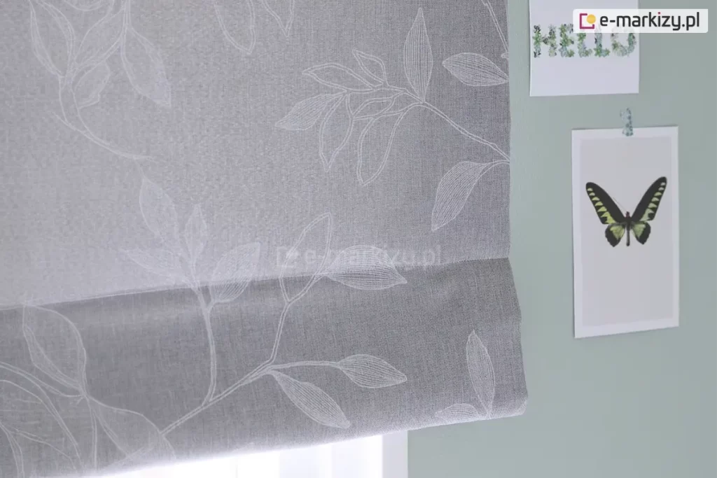 Zdjęcie przedstawia fragment dekoracyjnej rolety rzymskiej wykonanej z tkaniny w kolorze jasnoszarym o widocznym, gęstym splocie przypominającym strukturę lnu. Na powierzchni materiału znajduje się subtelny, biały wzór florystyczny przedstawiający kontury liści i gałązek. Linie tworzące motyw roślinny są delikatne i przypominają odręczny szkic lub haft, co nadaje osłonie okiennej lekkości i eleganckiego charakteru. Dolna część rolety jest podwinięta, tworząc charakterystyczną dla tego typu przesłon poziomą zakładkę, która podkreśla teksturę materiału. Roleta jest zamontowana na tle ściany pomalowanej na stonowany, szałwiowy lub jasnoturkusowy kolor. Po prawej stronie, obok krawędzi tkaniny, na ścianie przyklejone są dwie grafiki. Górna to podłużny kartonik z napisem HELLO ułożonym z motywów roślinnych, natomiast dolna to pionowa karta z centralnie umieszczoną grafiką przedstawiającą motyla o rozpostartych, ciemnozielonych skrzydłach z czarnymi i żółtymi akcentami. Całość kompozycji jest utrzymana w chłodnej, pastelowej kolorystyce, a oświetlenie padające od dołu sugeruje bliskość okna i naturalne źródło światła, które delikatnie przenika przez strukturę szarej tkaniny.