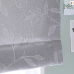 Zdjęcie przedstawia fragment dekoracyjnej rolety rzymskiej wykonanej z tkaniny w kolorze jasnoszarym o widocznym, gęstym splocie przypominającym strukturę lnu. Na powierzchni materiału znajduje się subtelny, biały wzór florystyczny przedstawiający kontury liści i gałązek. Linie tworzące motyw roślinny są delikatne i przypominają odręczny szkic lub haft, co nadaje osłonie okiennej lekkości i eleganckiego charakteru. Dolna część rolety jest podwinięta, tworząc charakterystyczną dla tego typu przesłon poziomą zakładkę, która podkreśla teksturę materiału. Roleta jest zamontowana na tle ściany pomalowanej na stonowany, szałwiowy lub jasnoturkusowy kolor. Po prawej stronie, obok krawędzi tkaniny, na ścianie przyklejone są dwie grafiki. Górna to podłużny kartonik z napisem HELLO ułożonym z motywów roślinnych, natomiast dolna to pionowa karta z centralnie umieszczoną grafiką przedstawiającą motyla o rozpostartych, ciemnozielonych skrzydłach z czarnymi i żółtymi akcentami. Całość kompozycji jest utrzymana w chłodnej, pastelowej kolorystyce, a oświetlenie padające od dołu sugeruje bliskość okna i naturalne źródło światła, które delikatnie przenika przez strukturę szarej tkaniny.