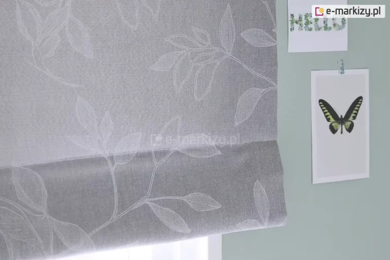 Zdjęcie przedstawia fragment dekoracyjnej rolety rzymskiej wykonanej z tkaniny w kolorze jasnoszarym o widocznym, gęstym splocie przypominającym strukturę lnu. Na powierzchni materiału znajduje się subtelny, biały wzór florystyczny przedstawiający kontury liści i gałązek. Linie tworzące motyw roślinny są delikatne i przypominają odręczny szkic lub haft, co nadaje osłonie okiennej lekkości i eleganckiego charakteru. Dolna część rolety jest podwinięta, tworząc charakterystyczną dla tego typu przesłon poziomą zakładkę, która podkreśla teksturę materiału. Roleta jest zamontowana na tle ściany pomalowanej na stonowany, szałwiowy lub jasnoturkusowy kolor. Po prawej stronie, obok krawędzi tkaniny, na ścianie przyklejone są dwie grafiki. Górna to podłużny kartonik z napisem HELLO ułożonym z motywów roślinnych, natomiast dolna to pionowa karta z centralnie umieszczoną grafiką przedstawiającą motyla o rozpostartych, ciemnozielonych skrzydłach z czarnymi i żółtymi akcentami. Całość kompozycji jest utrzymana w chłodnej, pastelowej kolorystyce, a oświetlenie padające od dołu sugeruje bliskość okna i naturalne źródło światła, które delikatnie przenika przez strukturę szarej tkaniny.