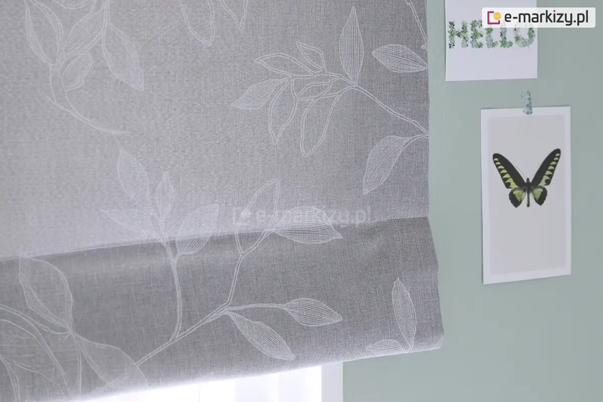 Zdjęcie przedstawia fragment dekoracyjnej rolety rzymskiej wykonanej z tkaniny w kolorze jasnoszarym o widocznym, gęstym splocie przypominającym strukturę lnu. Na powierzchni materiału znajduje się subtelny, biały wzór florystyczny przedstawiający kontury liści i gałązek. Linie tworzące motyw roślinny są delikatne i przypominają odręczny szkic lub haft, co nadaje osłonie okiennej lekkości i eleganckiego charakteru. Dolna część rolety jest podwinięta, tworząc charakterystyczną dla tego typu przesłon poziomą zakładkę, która podkreśla teksturę materiału. Roleta jest zamontowana na tle ściany pomalowanej na stonowany, szałwiowy lub jasnoturkusowy kolor. Po prawej stronie, obok krawędzi tkaniny, na ścianie przyklejone są dwie grafiki. Górna to podłużny kartonik z napisem HELLO ułożonym z motywów roślinnych, natomiast dolna to pionowa karta z centralnie umieszczoną grafiką przedstawiającą motyla o rozpostartych, ciemnozielonych skrzydłach z czarnymi i żółtymi akcentami. Całość kompozycji jest utrzymana w chłodnej, pastelowej kolorystyce, a oświetlenie padające od dołu sugeruje bliskość okna i naturalne źródło światła, które delikatnie przenika przez strukturę szarej tkaniny.