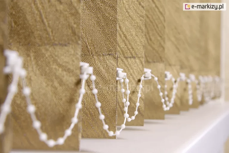 Zbliżenie na dolne wykończenie żaluzji pionowych typu vertikal wykonanych z dekoracyjnej tkaniny w odcieniu stonowanego złota lub musztardy o bogatej teksturze. Materiał posiada wyraźny, ukośny splot żakardowy tworzący wypukły wzór, który nadaje lamelom trójwymiarowy wygląd i elegancki charakter. Poszczególne pionowe pasy są ustawione pod kątem, a u ich dołu widoczne są białe, plastikowe uchwyty mocujące łańcuszek dolny. Biały łańcuszek koralikowy łączy każdą lamelę, tworząc powtarzalne, miękkie pętle, które stabilizują żaluzje pionowe vertikal i utrzymują je w równych odstępach. Tło obrazu jest celowo rozmyte, co skupia uwagę na detalach pierwszej lameli i precyzji mechanizmu łączącego. Całość kompozycji opiera się na jasnej, neutralnej powierzchni, prawdopodobnie parapecie, co podkreśla ciepłą kolorystykę osłony okiennej oraz jej techniczne detale wykończenia.