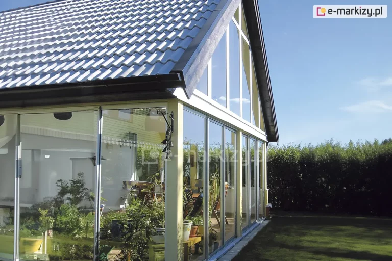 Zdjęcie przedstawia kompleksową zabudowę ogrodu zimowego lub wysokiego tarasu pod dachem dwuspadowym, wykonaną z wykorzystaniem systemu szklanych drzwi przesuwnych Sotro. Przeszklenie dolnej kondygnacji składa się z wielu paneli szklanych osadzonych w aluminiowych prowadnicach, które tworzą transparentną barierę wokół wnętrza wypełnionego roślinnością doniczkową i meblami ogrodowymi. Górna część zabudowy, wypełniająca trójkątny szczyt dachu, została wykonana z dopasowanych stałych naświetli szklanych w białych ramach, które harmonizują z konstrukcją budynku. Całość konstrukcji spoczywa na niskim fundamencie wyłożonym jasną kostką brukową, a szklane tafle odbijają błękit nieba oraz otaczającą ogród gęstą zieleń wysokiego żywopłotu. Architektura zabudowy pozwala na maksymalne doświetlenie wnętrza przy jednoczesnym zachowaniu pełnej ochrony przed wiatrem i deszczem, integrując dom z ogrodem.