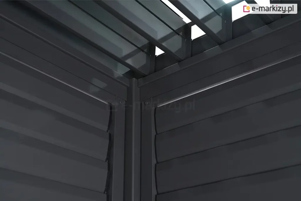 Zbliżenie na wewnętrzny narożnik konstrukcji pergoli tarasowej w kolorze antracytowym, przedstawiające punkt styku dwóch bocznych paneli żaluzjowych oraz zadaszenia lamelowego. W centralnej części widoczny jest pionowy profil narożny, do którego z obu stron przylegają ramy paneli bocznych wypełnione poziomymi, szerokimi lamelami. Lamele na ścianach bocznych są ustawione w pozycji zamkniętej lub lekko nachylonej, tworząc zwartą płaszczyznę o subtelnie wypukłym profilu. Powyżej ścian bocznych widoczny jest fragment konstrukcji dachu, w tym rynna wewnętrzna oraz mechanizm osadzenia lameli dachowych. Same lamele dachowe są ustawione prostopadle do belek konstrukcyjnych i znajdują się w pozycji otwartej, co pozwala na prześwit światła dziennego przez szczeliny między nimi. Całość wykonana jest z aluminium o matowym, fakturowanym wykończeniu. Perspektywa zdjęcia z dołu ku górze podkreśla masywność profili oraz precyzyjne dopasowanie elementów systemowych w narożniku budowli.