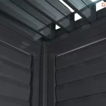 Zbliżenie na wewnętrzny narożnik konstrukcji pergoli tarasowej w kolorze antracytowym, przedstawiające punkt styku dwóch bocznych paneli żaluzjowych oraz zadaszenia lamelowego. W centralnej części widoczny jest pionowy profil narożny, do którego z obu stron przylegają ramy paneli bocznych wypełnione poziomymi, szerokimi lamelami. Lamele na ścianach bocznych są ustawione w pozycji zamkniętej lub lekko nachylonej, tworząc zwartą płaszczyznę o subtelnie wypukłym profilu. Powyżej ścian bocznych widoczny jest fragment konstrukcji dachu, w tym rynna wewnętrzna oraz mechanizm osadzenia lameli dachowych. Same lamele dachowe są ustawione prostopadle do belek konstrukcyjnych i znajdują się w pozycji otwartej, co pozwala na prześwit światła dziennego przez szczeliny między nimi. Całość wykonana jest z aluminium o matowym, fakturowanym wykończeniu. Perspektywa zdjęcia z dołu ku górze podkreśla masywność profili oraz precyzyjne dopasowanie elementów systemowych w narożniku budowli.