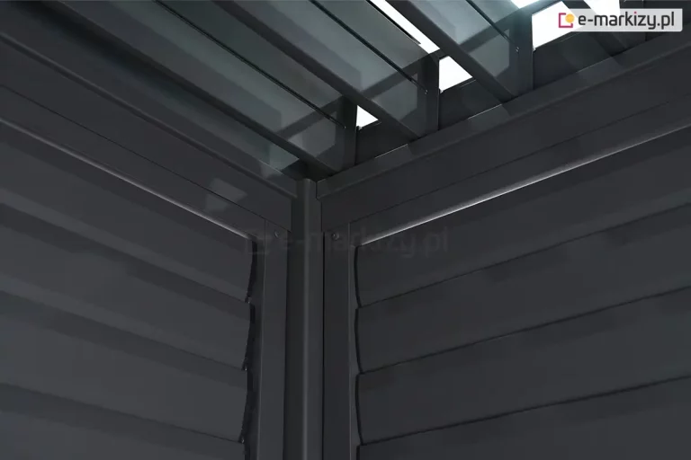 Zbliżenie na wewnętrzny narożnik konstrukcji pergoli tarasowej w kolorze antracytowym, przedstawiające punkt styku dwóch bocznych paneli żaluzjowych oraz zadaszenia lamelowego. W centralnej części widoczny jest pionowy profil narożny, do którego z obu stron przylegają ramy paneli bocznych wypełnione poziomymi, szerokimi lamelami. Lamele na ścianach bocznych są ustawione w pozycji zamkniętej lub lekko nachylonej, tworząc zwartą płaszczyznę o subtelnie wypukłym profilu. Powyżej ścian bocznych widoczny jest fragment konstrukcji dachu, w tym rynna wewnętrzna oraz mechanizm osadzenia lameli dachowych. Same lamele dachowe są ustawione prostopadle do belek konstrukcyjnych i znajdują się w pozycji otwartej, co pozwala na prześwit światła dziennego przez szczeliny między nimi. Całość wykonana jest z aluminium o matowym, fakturowanym wykończeniu. Perspektywa zdjęcia z dołu ku górze podkreśla masywność profili oraz precyzyjne dopasowanie elementów systemowych w narożniku budowli.