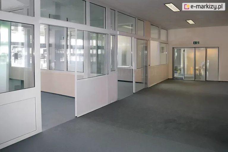 Przestronne wnętrze biurowe podzielone systemem ścianek stałych Fest, wykonanych z białych profili aluminiowych. Konstrukcja tworzy szereg przeszklonych pomieszczeń i boksów, które oddzielają strefy pracy od szerokiego ciągu komunikacyjnego. Poszczególne moduły ścianek posiadają mieszane wypełnienie: dolne sekcje wykonano z nieprzeziernych paneli w kolorze białym, natomiast górne partie są w całości przeszklone, co zapewnia swobodny przepływ światła i optyczną przestronność. W systemie zintegrowano przesuwne drzwi wejściowe do poszczególnych biur, osadzone w aluminiowych prowadnicach. Na podłodze korytarza oraz wewnątrz boksów znajduje się jednolita, ciemnoszara wykładzina obiektowa o krótkim włosiu. W głębi korytarza widoczne jest przeszklone wyjście ewakuacyjne oznaczone zielonym piktogramem, a na suficie zamontowano podłużne lampy jarzeniowe zapewniające równomierne oświetlenie techniczne. Całość aranżacji utrzymana jest w profesjonalnej, biało-szarej kolorystyce, podkreślającej sterylny i nowoczesny charakter biura.