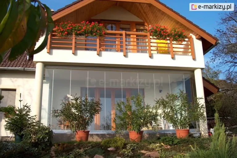 Parter domu jednorodzinnego z pełną, bezramową zabudową szklaną tarasu Miros. Przezroczyste panele sięgają od podłogi do sufitu, tworząc lekką, nowoczesną fasadę, za którą widoczna jest drewniana stolarka drzwiowa oraz białe krzesła. Nad przeszkleniem znajduje się balkon z drewnianą balustradą i skrzynkami z czerwonymi pelargoniami. Dach budynku dwuspadowy, z drewnianym wykończeniem podbitki. Przed przeszklonym tarasem ustawiono donice z dużymi roślinami ozdobnymi.