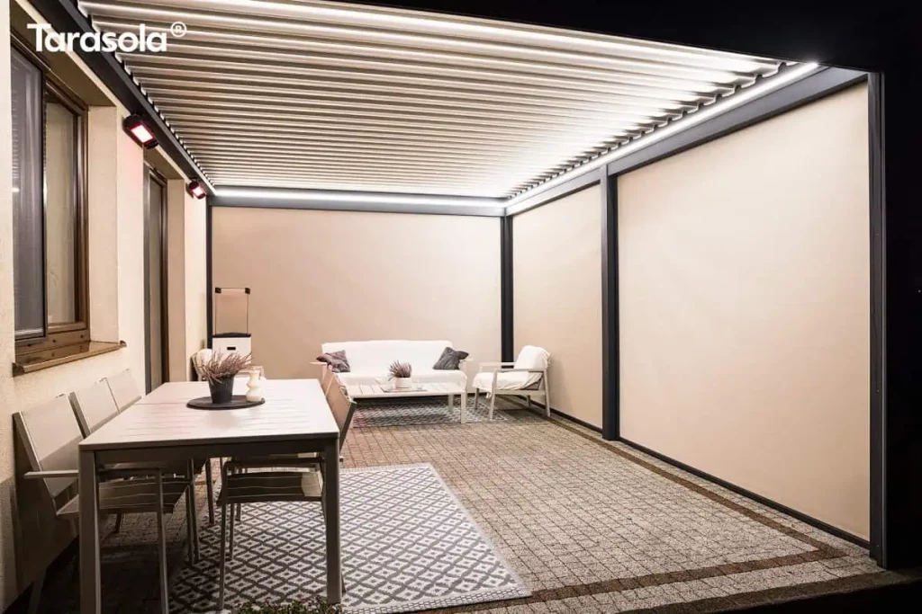 Nowoczesna pergola bioklimatyczna essential Tarasola w kolorze ciemnoantracytowym, przedstawiona od wewnątrz w warunkach wieczornych. Konstrukcja posiada dach z ruchomych lameli oraz w pełni opuszczone boczne rolety typu screen w kolorze beżowym, które szczelnie zamykają przestrzeń, zapewniając prywatność i ochronę przed wiatrem. Wnętrze jest intensywnie oświetlone za pomocą liniowych listew LED zamontowanych na obwodzie ramy dachowej, a na ścianie budynku widoczne są dodatkowe promienniki ciepła emitujące czerwone światło. Pod zadaszeniem znajduje się strefa jadalniana z szarym stołem i krzesłami oraz strefa wypoczynkowa z białą sofą, fotelami i stolikiem kawowym, ustawionymi na podłożu z kostki brukowej z ozdobnymi dywanami zewnętrznymi. Zdjęcie podkreśla funkcjonalność osłon bocznych i systemów grzewczych, które pozwalają na komfortowe korzystanie z tarasu po zmroku.