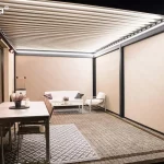 Nowoczesna pergola bioklimatyczna essential Tarasola w kolorze ciemnoantracytowym, przedstawiona od wewnątrz w warunkach wieczornych. Konstrukcja posiada dach z ruchomych lameli oraz w pełni opuszczone boczne rolety typu screen w kolorze beżowym, które szczelnie zamykają przestrzeń, zapewniając prywatność i ochronę przed wiatrem. Wnętrze jest intensywnie oświetlone za pomocą liniowych listew LED zamontowanych na obwodzie ramy dachowej, a na ścianie budynku widoczne są dodatkowe promienniki ciepła emitujące czerwone światło. Pod zadaszeniem znajduje się strefa jadalniana z szarym stołem i krzesłami oraz strefa wypoczynkowa z białą sofą, fotelami i stolikiem kawowym, ustawionymi na podłożu z kostki brukowej z ozdobnymi dywanami zewnętrznymi. Zdjęcie podkreśla funkcjonalność osłon bocznych i systemów grzewczych, które pozwalają na komfortowe korzystanie z tarasu po zmroku.