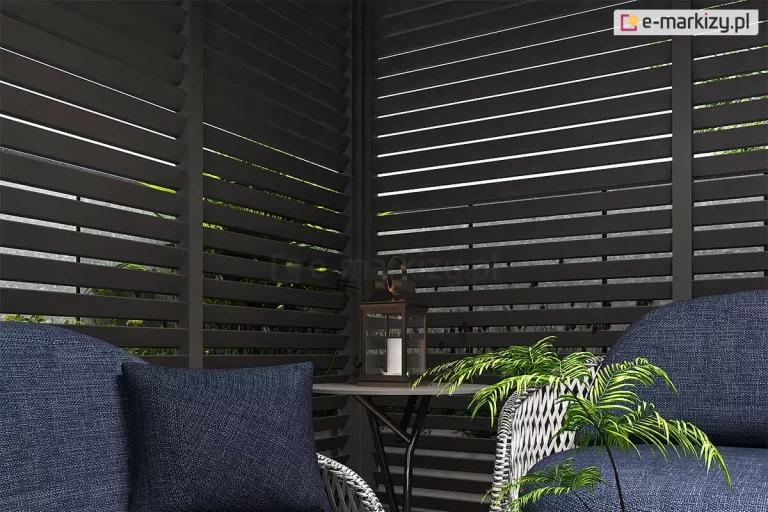 Wnętrze pergoli Marlo wyposażonej w narożną zabudowę z antracytowych paneli żaluzjowych, tworzących intymną strefę wypoczynkową. Poziome, nieruchome lamele paneli bocznych są ustawione pod stałym kątem, co zapewnia prywatność oraz ochronę przed słońcem, jednocześnie umożliwiając subtelną cyrkulację powietrza. Wewnątrz strefy znajdują się meble ogrodowe z ciemnoniebieskimi poduszkami o wyraźnej strukturze splotu, ustawione obok okrągłego stolika pomocniczego o cienkim blacie. Na stoliku umieszczono klasyczną latarnię ogrodową z białą świecą wewnątrz. W tle, pomiędzy szczelinami żaluzji, widoczne są fragmenty roślinności ogrodowej. Zdjęcie wykonane z bliskiej perspektywy podkreśla matowe wykończenie profili aluminiowych oraz detale wykończenia tekstyliów meblowych.