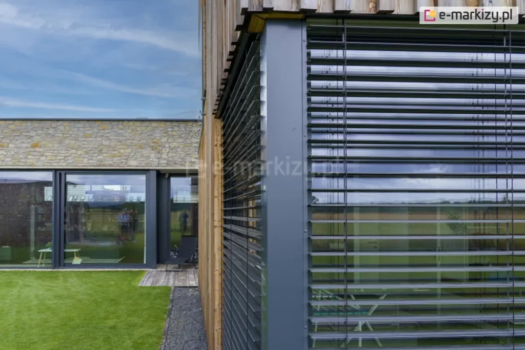 Zbliżenie na róg nowoczesnego domu z dużymi przeszkleniami zabezpieczonymi zewnętrznymi żaluzjami fasadowymi. System żaluzji wykonany z poziomych lameli aluminiowych, zamontowany na antracytowych prowadnicach w elewacji wykończonej deską elewacyjną. Obok znajduje się fragment budynku z elewacją z naturalnego kamienia. Na parterze przeszklone ściany i drzwi tarasowe, w których odbija się niebo, trawnik oraz sylwetki osób. W dolnej części widoczny zielony trawnik oraz pas ciemnego żwiru przy budynku.