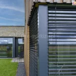 Zbliżenie na róg nowoczesnego domu z dużymi przeszkleniami zabezpieczonymi zewnętrznymi żaluzjami fasadowymi. System żaluzji wykonany z poziomych lameli aluminiowych, zamontowany na antracytowych prowadnicach w elewacji wykończonej deską elewacyjną. Obok znajduje się fragment budynku z elewacją z naturalnego kamienia. Na parterze przeszklone ściany i drzwi tarasowe, w których odbija się niebo, trawnik oraz sylwetki osób. W dolnej części widoczny zielony trawnik oraz pas ciemnego żwiru przy budynku.