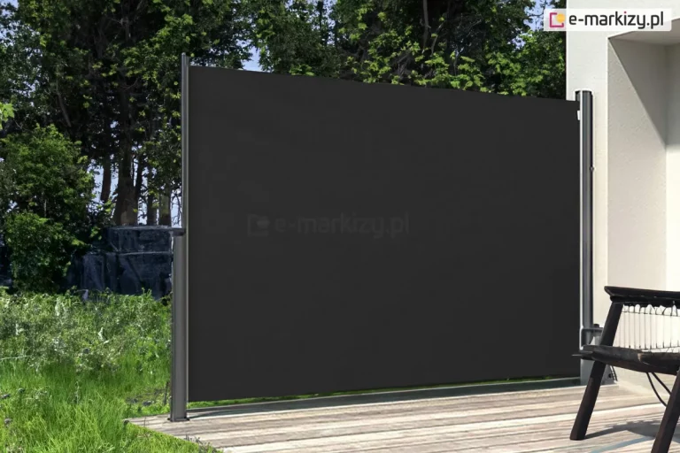 Zdjęcie prezentuje boczną markizę tarasową w formie rozwiniętego ekranu, zainstalowaną na nowoczesnym tarasie wyłożonym deską kompozytową w jasnym kolorze z wyraźnym usłojeniem. Przesłona składa się z pionowej, srebrnej kasety zamontowanej do jasnej, otynkowanej ściany budynku oraz z tkaniny w kolorze głębokiej czerni, która jest całkowicie rozciągnięta. Materiał jest zamocowany do wolnostojącego, ciemnoszarego słupka końcowego przytwierdzonego do podłoża za pomocą stopy montażowej. Tkanina markizy jest napięta, tworząc jednolitą, nieprzezierną barierę chroniącą przed słońcem, wiatrem oraz zapewniającą prywatność. Po prawej stronie kadru widoczny jest fragment ciemnego, drewnianego krzesła ogrodowego o ażurowej konstrukcji siedziska. W tle, za markizą, rozpościera się widok na ogród z gęstą ścianą zielonych drzew i krzewów oraz fragmentem trawiastego podłoża. Scena jest oświetlona naturalnym światłem dziennym, które podkreśla kontrast między czarnym materiałem osłony a jasną elewacją i podłogą tarasu.