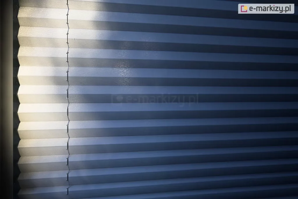 Zdjęcie prezentuje bliskie ujęcie na rozwiniętą osłonę okienną, którą jest żaluzja plisowana w chłodnym, niebieskoszarym lub stalowym odcieniu. Plisa na okno charakteryzuje się gęstym, poziomym żebrowaniem, tworzącym rytmiczny układ załamań materiału. Lewa strona tkaniny jest intensywnie oświetlona silnym strumieniem światła padającym z boku, co sprawia, że krawędzie plis stają się niemal białe i zyskują lekko błyszczącą, ziarnistą fakturę. Prawa część zdjęcia pozostaje w głębokim cieniu, co podkreśla głębię koloru i trójwymiarowość konstrukcji. Przez lewą część materiału przebiega pionowa linia sznurka prowadzącego, który jest precyzyjnie przewleczony przez system małych otworów w każdym zagięciu harmonijki. Gra światła i cienia na powierzchni tkaniny uwidacznia strukturę materiału oraz techniczny aspekt prowadzenia linek stabilizujących. Kadr jest ciasny, skupiony wyłącznie na teksturze i geometrii żaluzji, bez widocznych elementów ramy czy otoczenia.
