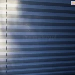 Zdjęcie prezentuje bliskie ujęcie na rozwiniętą osłonę okienną, którą jest żaluzja plisowana w chłodnym, niebieskoszarym lub stalowym odcieniu. Plisa na okno charakteryzuje się gęstym, poziomym żebrowaniem, tworzącym rytmiczny układ załamań materiału. Lewa strona tkaniny jest intensywnie oświetlona silnym strumieniem światła padającym z boku, co sprawia, że krawędzie plis stają się niemal białe i zyskują lekko błyszczącą, ziarnistą fakturę. Prawa część zdjęcia pozostaje w głębokim cieniu, co podkreśla głębię koloru i trójwymiarowość konstrukcji. Przez lewą część materiału przebiega pionowa linia sznurka prowadzącego, który jest precyzyjnie przewleczony przez system małych otworów w każdym zagięciu harmonijki. Gra światła i cienia na powierzchni tkaniny uwidacznia strukturę materiału oraz techniczny aspekt prowadzenia linek stabilizujących. Kadr jest ciasny, skupiony wyłącznie na teksturze i geometrii żaluzji, bez widocznych elementów ramy czy otoczenia.