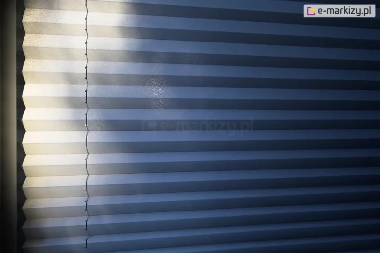 Zdjęcie prezentuje bliskie ujęcie na rozwiniętą osłonę okienną, którą jest żaluzja plisowana w chłodnym, niebieskoszarym lub stalowym odcieniu. Plisa na okno charakteryzuje się gęstym, poziomym żebrowaniem, tworzącym rytmiczny układ załamań materiału. Lewa strona tkaniny jest intensywnie oświetlona silnym strumieniem światła padającym z boku, co sprawia, że krawędzie plis stają się niemal białe i zyskują lekko błyszczącą, ziarnistą fakturę. Prawa część zdjęcia pozostaje w głębokim cieniu, co podkreśla głębię koloru i trójwymiarowość konstrukcji. Przez lewą część materiału przebiega pionowa linia sznurka prowadzącego, który jest precyzyjnie przewleczony przez system małych otworów w każdym zagięciu harmonijki. Gra światła i cienia na powierzchni tkaniny uwidacznia strukturę materiału oraz techniczny aspekt prowadzenia linek stabilizujących. Kadr jest ciasny, skupiony wyłącznie na teksturze i geometrii żaluzji, bez widocznych elementów ramy czy otoczenia.