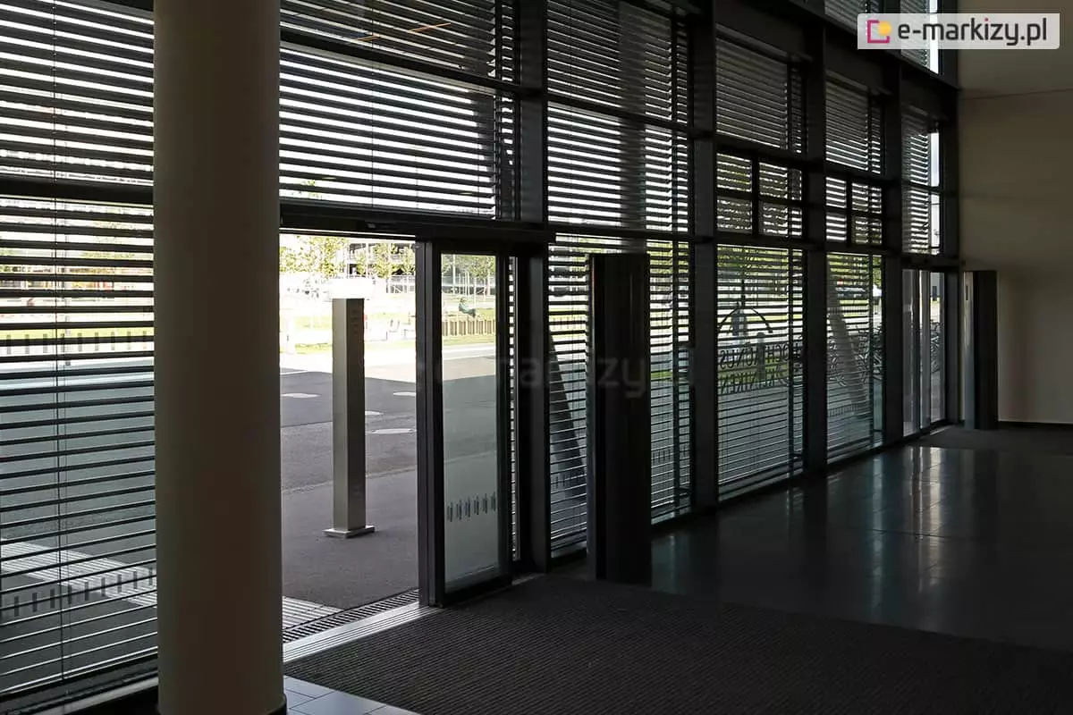 Widok z wnętrza nowoczesnego holu budynku na przeszkloną ścianę frontową z zainstalowanymi żaluzjami fasadowymi Selt. Poziome lamele żaluzji są ustawione w pozycji półotwartej, co pozwala na przenikanie światła dziennego do środka, tworząc rytmiczny wzór cieni na podłodze, jednocześnie ograniczając nagrzewanie pomieszczenia. Przez przeszklenia widoczny jest plac zewnętrzny oraz fragmenty zieleni miejskiej. Konstrukcja żaluzji obejmuje całą wysokość witryn, od podłogi aż po sufit, z wyraźnie zaznaczonymi podziałami pionowymi ram okiennych i drzwi wejściowych. Po lewej stronie kadru widoczny jest masywny filar konstrukcyjny budynku, a wewnątrz znajdują się elementy infrastruktury, takie jak ciemna wycieraczka systemowa i metalowe słupki informacyjne.
