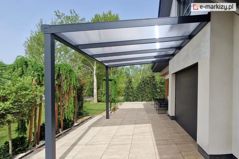 Boczne ujęcie nowoczesnego zadaszenia tarasowego Gumax (obecnie Deponti) o konstrukcji w kolorze antracytowym, które zostało zamontowane do jasnej elewacji piętrowego domu. Aluminiowa pergola posiada jednospadowy dach z wypełnieniem z mlecznych, półprzezroczystych paneli poliwęglanowych, wsparty na dwóch solidnych, kwadratowych słupach nośnych. Pod zadaszeniem znajduje się przestronny taras wyłożony beżowymi płytami o teksturze drewna, na którym w głębi ustawiono czarną, metalową ławkę. Na ścianie budynku widoczna jest duża, ciemnoszara roleta zewnętrzna oraz ozdobny fragment elewacji z drewnopodobnych paneli. Otoczenie stanowi gęsty ogród z wysokimi brzozami, krzewami oraz fragmentem iglaka na pierwszym planie, a całość dopełnia bezchmurne, jasne niebo.