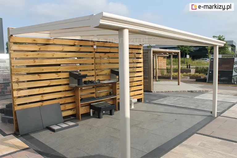 Niestandardowe zastosowanie nowoczesnego zadaszenia tarasowego Lunaro w kolorze kremowym. Konstrukcja o klasycznym wykończeniu i półokrągłych słupach frontowych zamocowana została do konstrukcji, wykończonej drewnianymi paletami. Zadaszenie zostało ustawione na ekspozycji zewnętrznej wyłożonej dużymi, ciemnoszarymi płytami tarasowymi, a jego tylną część ogranicza ażurowa ściana wykonana z poziomych, drewnianych desek o naturalnym odcieniu. Na regale przy drewnianej ścianie oraz na podłożu wyeksponowano różne akcesoria montażowe, próbki materiałów oraz elementy systemu odwodnienia. W tle widoczne są inne konstrukcje ogrodowe, fragmenty brukowanych alejek oraz budynek handlowy pod jasnym, lekko zachmurzonym niebem.