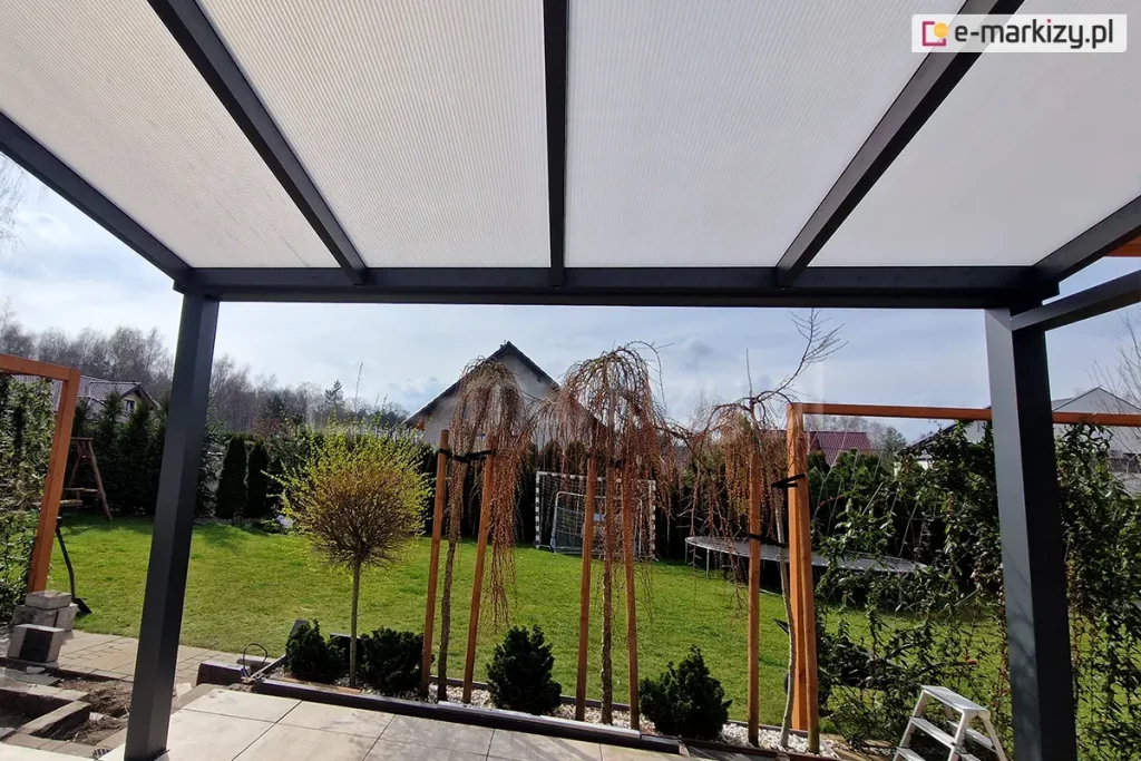 Zadaszenie z panelami poliwęglanowymi w kolorze opalowym oraz antracytową konstrukcją, ustawione nad tarasem z płyt gresowych. Na krawędzi tarasu widoczne drzewka bez liści oraz niskie iglaki posadzone w donicach. W tle widoczny trawnik z bramkami do gry w piłkę oraz dużą trampoliną, otoczony żywopłotem z tui, nad którym rozciąga się lekko zachmurzone szare niebo.