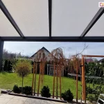 Zadaszenie z panelami poliwęglanowymi w kolorze opalowym oraz antracytową konstrukcją, ustawione nad tarasem z płyt gresowych. Na krawędzi tarasu widoczne drzewka bez liści oraz niskie iglaki posadzone w donicach. W tle widoczny trawnik z bramkami do gry w piłkę oraz dużą trampoliną, otoczony żywopłotem z tui, nad którym rozciąga się lekko zachmurzone szare niebo.