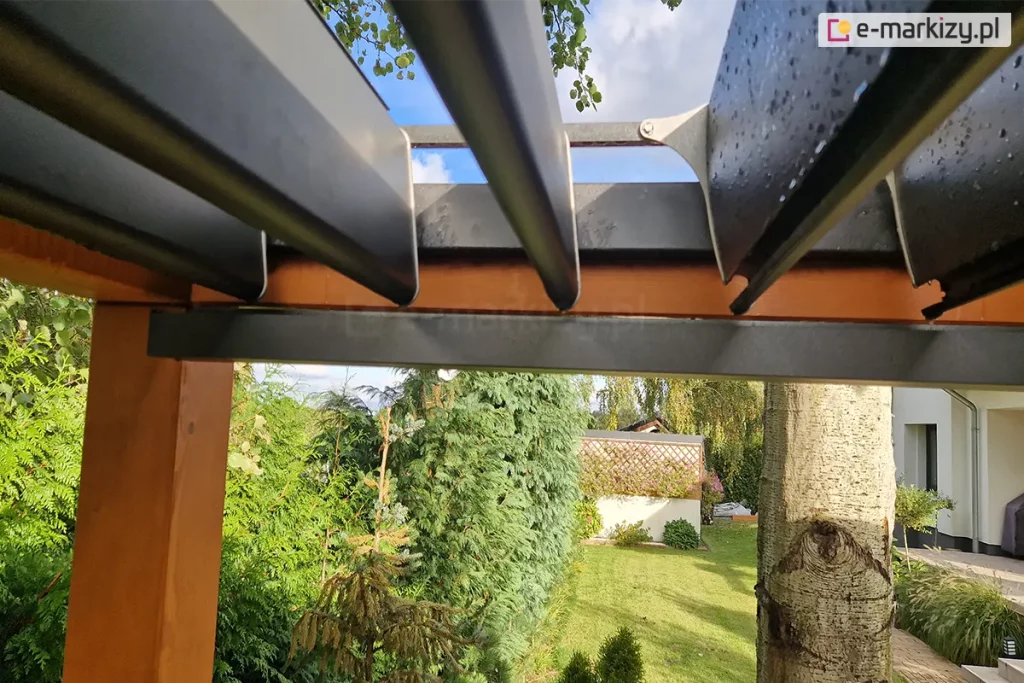 Moduł dachowy SB400 Selt w kolorze antracytowym zamocowany na konstrukcji drewnianej pergoli. Uchylone lamele pokryte kroplami wody ukazują niebieskie, rozsłonecznione niebo. Pod pergolą widoczny ogród z trawnikiem narożnik budynku oraz liczne drzewa i krzewy.