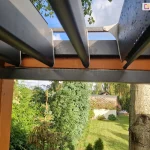 Moduł dachowy SB400 Selt w kolorze antracytowym zamocowany na konstrukcji drewnianej pergoli. Uchylone lamele pokryte kroplami wody ukazują niebieskie, rozsłonecznione niebo. Pod pergolą widoczny ogród z trawnikiem narożnik budynku oraz liczne drzewa i krzewy.