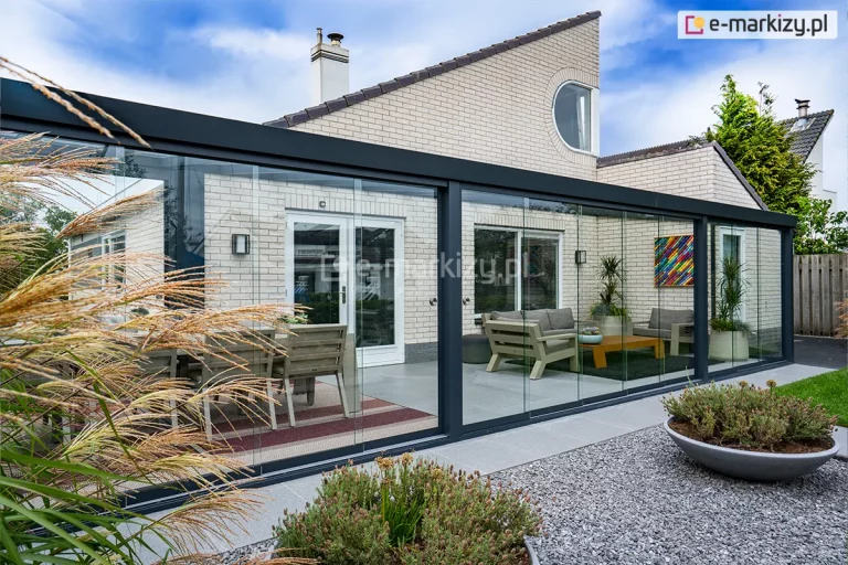 Nowoczesna realizacja całorocznej zabudowy tarasu, zintegrowanej z bryłą budynku jednorodzinnego o jasnej elewacji. Konstrukcja nośna została wykonana z grubych profili aluminiowych w kolorze antracytowym, które tworzą geometryczną ramę zadaszenia oraz ścian bocznych. W systemie wykorzystano szklane ściany przesuwne, które dzielą przestrzeń na regularne sekcje, zapewniając pełną przezierność i ochronę przed warunkami atmosferycznymi. Dach konstrukcji jest w całości przeszklony, co pozwala na maksymalne doświetlenie wnętrza światłem dziennym. Wewnątrz przeszklonej strefy zaaranżowano dwie przestrzenie: jadalnianą ze stołem i drewnianymi krzesłami oraz wypoczynkową z fotelami i niskim stolikiem kawowym. Podłoże tarasu stanowią wielkoformatowe płyty w kolorze jasnoszarym, które płynnie łączą się z obszarem wysypanym szarym gresem. Całość kompozycji dopełnia niska roślinność ogrodowa w ozdobnych donicach oraz ozdobne trawy na pierwszym planie, co podkreśla spójność nowoczesnej architektury z naturą.