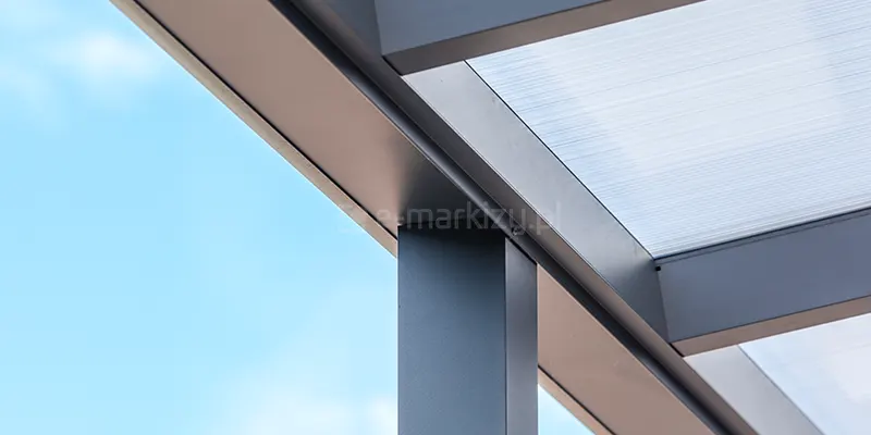 zbliżenie na górny narożnik konstrukcji aluminiowego zadaszenia tarasowego w kolorze antracytowym. Widoczny jest punkt styku poziomej belki rynnowej z pionowym słupem nośnym o kwadratowym przekroju oraz prostopadle osadzoną krokwią dachową. Na krokwiach spoczywa płyta z przezroczystego poliwęglanu komorowego, którego liniowa struktura wewnętrzna jest wyraźnie widoczna pod kątem. Profile posiadają gładkie, matowe wykończenie i są precyzyjnie spasowane. W tle widoczne jest jasnobłękitne, bezchmurne niebo, co podkreśla detale techniczne górnej części systemu zadaszenia.