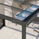 Zadaszenie tarasu ze szkła Terano z klasycznym wykończeniem belki przedniej, w której ukryta jest rynna. Pod antracytową konstrukcją zadaszenia oraz przejrzystym szkłem widoczny taras wyłożony dużymi jasnymi płytkami oraz zestaw mebli z rattanu z poduchami w kolorach szarym i zielonym, a także niski stolik kawowy w odcieniu grafitowym.