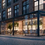 Zdjęcie prezentuje wieczorną wizualizację nowoczesnego, w pełni przeszklonego zadaszenia Lunaro, zaadaptowanego na potrzeby ogródka restauracyjnego przy budynku z czerwonej cegły. Konstrukcja w kolorze antracytowym posiada szklane ściany przesuwne, które ukazują eleganckie wnętrze z drewnianymi stolikami, plecionymi fotelami oraz pnącą roślinnością na ścianach. Ciepłe, punktowe oświetlenie zamontowane w krokwiach dachowych oraz neonowy napis wewnątrz lokalu tworzą przytulną atmosferę. Całość znajduje się przy brukowanej ulicy, a przed wejściem ustawiono symetrycznie małe drzewka w ozdobnych białych donicach.