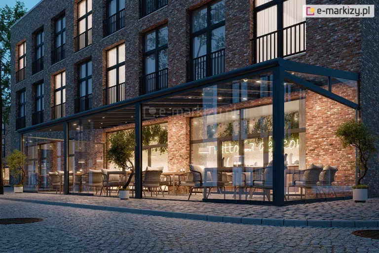Zdjęcie prezentuje wieczorną wizualizację nowoczesnego, w pełni przeszklonego zadaszenia Lunaro, zaadaptowanego na potrzeby ogródka restauracyjnego przy budynku z czerwonej cegły. Konstrukcja w kolorze antracytowym posiada szklane ściany przesuwne, które ukazują eleganckie wnętrze z drewnianymi stolikami, plecionymi fotelami oraz pnącą roślinnością na ścianach. Ciepłe, punktowe oświetlenie zamontowane w krokwiach dachowych oraz neonowy napis wewnątrz lokalu tworzą przytulną atmosferę. Całość znajduje się przy brukowanej ulicy, a przed wejściem ustawiono symetrycznie małe drzewka w ozdobnych białych donicach.