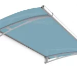 Zdjęcie obrazuje zadaszenie drzwiowe Elegant z barwionym wypełnieniem akrylowym w kolorze jasnoniebieskim o lekko dymionym stopniu przezroczystości. Niebieska płyta jest osadzona na dwóch aluminiowych wspornikach w kolorze srebrnym, które mają łukowaty kształt i ażurowe zdobienia. Przednia krawędź daszka wyposażona jest w rynnę odpływową o okrągłym profilu, która ułatwia odprowadzanie deszczówki na boki. Konstrukcja jest nachylona pod kątem, a jej tylna listwa montażowa zapewnia stabilne przyleganie do płaszczyzny ściany. Kolorystyka zadaszenia łączy chłodny odcień błękitu z metalicznymi, stalowymi elementami stelaża.