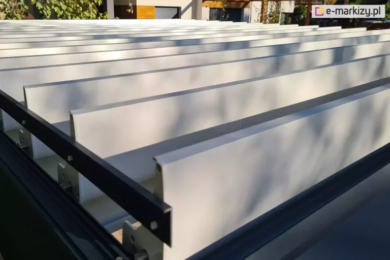 Widok z bliska na górną strukturę zadaszenia nowoczesnej pergoli bioklimatycznej, prezentujący rzędy aluminiowych lamel w kolorze białym. Lamele są ustawione w pozycji otwartej, niemal pionowo względem ramy, co pozwala na pełną wentylację i dopływ światła. Każdy z białych profili dachowych posiada precyzyjnie wykończone krawędzie i jest zamocowany do bocznej, czarnej szyny napędowej za pomocą widocznych stalowych sworzni i elementów montażowych, które stanowią oś obrotu dla mechanizmu regulacji kąta nachylenia. Rama zewnętrzna oraz szyna prowadząca zostały wykonane z aluminium lakierowanego proszkowo na głęboki, matowy odcień antracytu. Na gładkiej powierzchni białych lamel widoczne są miękkie, plastyczne cienie rzucane przez liście pobliskich drzew, co świadczy o wysokiej jakości wykończenia powłoki lakierniczej. W rozmytym tle, poza obszarem ostrości, dostrzec można fragmenty nowoczesnej elewacji budynku mieszkalnego oraz naturalną zieleń ogrodową. Perspektywa zdjęcia pod kątem od góry akcentuje rytmiczny układ elementów konstrukcyjnych oraz techniczną precyzję systemu sterowania zadaszeniem lamelowym.