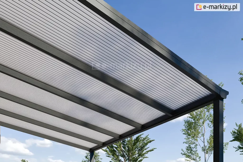 Perspektywiczny widok od dołu na narożnik nowoczesnego zadaszenia tarasowego Lunaro, wykonanego z antracytowych profili aluminiowych. Konstrukcja dachu składa się z grubych, równoległych krokwi, pomiędzy którymi zamontowano panele z poliwęglanu komorowego w kolorze opalowym (mlecznym). Półprzezroczyste wypełnienie dachu charakteryzuje się wyraźną strukturą pionowych linii, która skutecznie rozprasza światło słoneczne. W tle, pod czystym błękitnym niebem, widoczne są wierzchołki zielonych drzew, co podkreśla ogrodowy charakter instalacji.