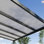 Perspektywiczny widok od dołu na narożnik nowoczesnego zadaszenia tarasowego Lunaro, wykonanego z antracytowych profili aluminiowych. Konstrukcja dachu składa się z grubych, równoległych krokwi, pomiędzy którymi zamontowano panele z poliwęglanu komorowego w kolorze opalowym (mlecznym). Półprzezroczyste wypełnienie dachu charakteryzuje się wyraźną strukturą pionowych linii, która skutecznie rozprasza światło słoneczne. W tle, pod czystym błękitnym niebem, widoczne są wierzchołki zielonych drzew, co podkreśla ogrodowy charakter instalacji.