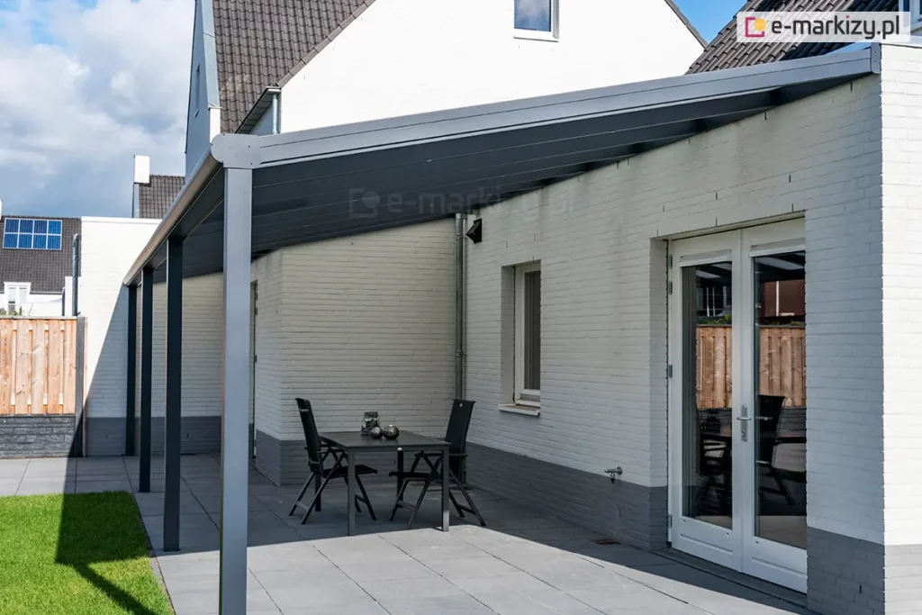 Nowoczesne zadaszenie tarasowe Bosco Deponti (nazwa handlowa Lunaro), opartego na czterech solidnych słupach nośnych, zamontowanego do elewacji białego budynku z cegły. Konstrukcja w kolorze ciemnego antracytu posiada duży, jednospadowy dach z wypełnieniem z poliwęglanu, który osłania przestronny taras wyłożony szarymi płytami. Pod zadaszeniem ustawiono prosty, czarny stół z dwoma dopasowanymi krzesłami, a w tle widoczne są białe, przeszklone drzwi tarasowe prowadzące do wnętrza domu. Na dalszym planie znajduje się fragment zielonego trawnika oraz drewniane ogrodzenie, nad którymi góruje biała ściana szczytowa budynku z małym oknem pod dachem z ciemnej dachówki.