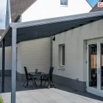 Nowoczesne zadaszenie tarasowe Bosco Deponti (nazwa handlowa Lunaro), opartego na czterech solidnych słupach nośnych, zamontowanego do elewacji białego budynku z cegły. Konstrukcja w kolorze ciemnego antracytu posiada duży, jednospadowy dach z wypełnieniem z poliwęglanu, który osłania przestronny taras wyłożony szarymi płytami. Pod zadaszeniem ustawiono prosty, czarny stół z dwoma dopasowanymi krzesłami, a w tle widoczne są białe, przeszklone drzwi tarasowe prowadzące do wnętrza domu. Na dalszym planie znajduje się fragment zielonego trawnika oraz drewniane ogrodzenie, nad którymi góruje biała ściana szczytowa budynku z małym oknem pod dachem z ciemnej dachówki.