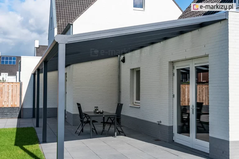 Nowoczesne zadaszenie tarasowe Bosco Deponti (nazwa handlowa Lunaro), opartego na czterech solidnych słupach nośnych, zamontowanego do elewacji białego budynku z cegły. Konstrukcja w kolorze ciemnego antracytu posiada duży, jednospadowy dach z wypełnieniem z poliwęglanu, który osłania przestronny taras wyłożony szarymi płytami. Pod zadaszeniem ustawiono prosty, czarny stół z dwoma dopasowanymi krzesłami, a w tle widoczne są białe, przeszklone drzwi tarasowe prowadzące do wnętrza domu. Na dalszym planie znajduje się fragment zielonego trawnika oraz drewniane ogrodzenie, nad którymi góruje biała ściana szczytowa budynku z małym oknem pod dachem z ciemnej dachówki.