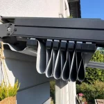Zdjęcie prezentuje techniczne zbliżenie na system zadaszenia tarasowego Square zamontowany do elewacji budynku. Głównym elementem kadru jest masywna, antracytowa prowadnica boczna o matowym wykończeniu, pod którą znajduje się mechanizm przesuwny. Tkanina zadaszenia w kolorze jasnoszarym jest zwinięta w pakiet, tworząc serię pionowych, regularnych fałd o zaokrąglonych krawędziach. Poszczególne sekcje materiału są zamocowane do czarnych, systemowych wózków jezdnych, które poruszają się wewnątrz szyny. Konstrukcja jest przytwierdzona do jasnej ściany budynku za pomocą ściennych uchwytów montażowych z widocznymi śrubami. W tle, pod zadaszeniem, widać fragment tarasu z meblami ogrodowymi, donice z roślinami oraz gęstą zieleń ogrodową. Oświetlenie jest ostre i słoneczne, co podkreśla kontrast między ciemnym profilem konstrukcyjnym a jasnym poszyciem materiałowym.