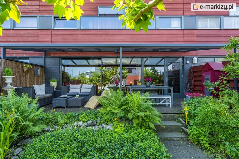 Zdjęcie prezentuje rozbudowaną strefę wypoczynkową na tarasie, osłoniętą szerokim zadaszeniem Nebbiolo marki Deponti. Konstrukcja z aluminium w kolorze antracytowym rozciąga się wzdłuż elewacji nowoczesnego budynku, która łączy ciemnoszarą cegłę na parterze z czerwonymi panelami drewnianymi na piętrze. Pod zadaszeniem, na drewnianym podeście, zaaranżowano dwie funkcjonalne przestrzenie: jadalnianą z białym stołem i ławami oraz wypoczynkową z grafitowymi meblami technorattanowymi i szarymi poduszkami. Duże przeszklenia parteru odbijają otaczającą zieleń, a boczna część tarasu została zabezpieczona szklanym klinem pod dachem oraz matą osłonową na ogrodzeniu. Pierwszy plan zajmuje bogata roślinność ogrodowa, w tym rozłożyste paprocie, funkie oraz krzewy, a w rogu widoczny jest dekoracyjny, mały czerwony domek narzędziowy.