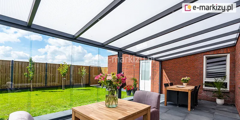 Perspektywa z wnętrza nowocześnie zabudowanego tarasu, zrealizowanego przy budynku z czerwonej cegły. Konstrukcja zadaszenia wykonana jest z aluminiowych profili w kolorze antracytowym, a dach wypełniają płyty z mlecznego poliwęglanu komorowego. Przestrzeń tarasu została zamknięta systemem bezramowych, przesuwnych ścian szklanych Auris, które oferują panoramiczny widok na ogród. Wewnątrz znajdują się dwie strefy: na pierwszym planie drewniany stolik kawowy z wazonem kolorowych kwiatów i szarym fotelem, a w głębi strefa jadalniana z większym stołem i czarnymi krzesłami z technorattanu. Posadzka wykonana jest z dużych, grafitowych płyt tarasowych. W tle, za szklaną zabudową, widoczny jest zielony trawnik, młode drzewka oraz wysokie drewniane ogrodzenie pod błękitnym niebem z rozproszonymi chmurami.