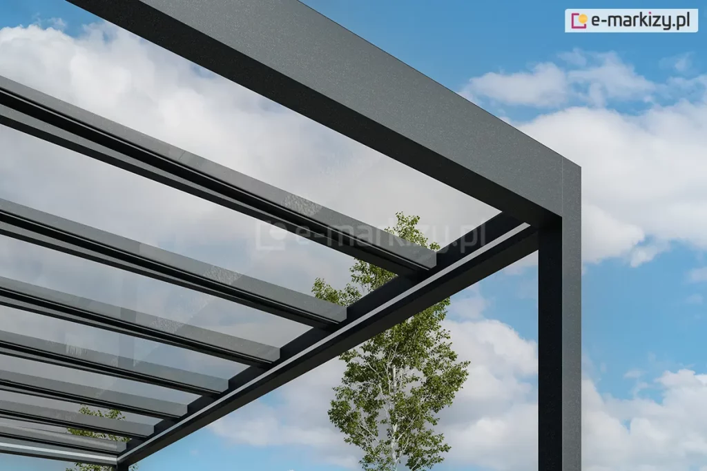 Zbliżenie na górny narożnik i konstrukcję dachu nowoczesnego zadaszenia tarasowego Spekta Glass, ukazane na tle błękitnego nieba z rozproszonymi białymi chmurami. Konstrukcja nośna została wykonana z solidnych profili aluminiowych w kolorze antracytowym o subtelnej, matowej teksturze, tworzących geometryczną ramę wspartą na pionowym słupie. Dach zadaszenia składa się z równolegle rozmieszczonych krokwi aluminiowych, na których zamontowano tafle w pełni przeziernego, bezpiecznego szkła. System szklanych paneli zapewnia maksymalny dostęp światła dziennego do przestrzeni pod zadaszeniem, jednocześnie chroniąc ją przed opadami. Pomiędzy profilami dachu widoczna jest korona zielonego drzewa, co podkreśla minimalistyczny design konstrukcji, która harmonijnie wtapia się w otoczenie. Całość prezentuje techniczny detal łączenia profili oraz wysoką jakość wykończenia elementów systemowych.